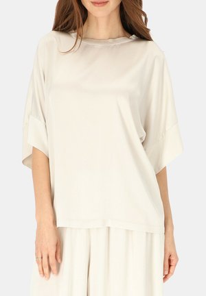 Kobieta w luźnej, kremowej satynowej bluzce z krótkim rękawem oraz pasujących szerokich spodniach, stojąca na jednolitym tle.