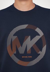 Sweatshirt azul marinho com um logotipo circular apresentando as iniciais listradas "MK" em branco e laranja, com o texto "MICHAEL KORS" abaixo.