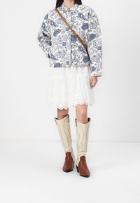 Pikowana kurtka z niebieskim wzorem kwiatowym, kremowe tło; warstwowa biała spódnica z ażurowego materiału; beżowe kowbojki z brązowymi akcentami.