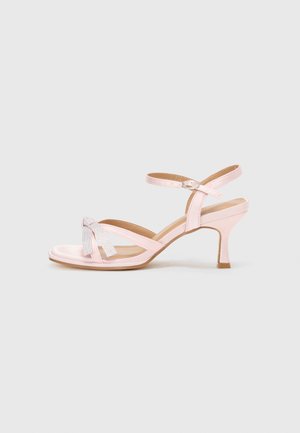 Sandales à talons roses avec finition brillante, présentant des brides croisées et un nœud décoratif en matériau métallique. Bride de cheville ajustable.