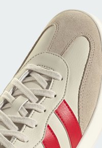 Baskets beige avec bout en daim, lacets blancs et deux bandes en cuir rouge sur le côté, présentées en gros plan.