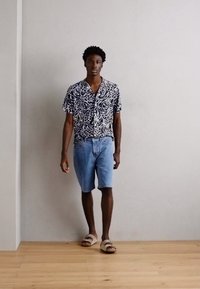 Camisa de mangas curtas azul-marinho e branca com padrão, calções de ganga azul claro e sandálias bege num modelo contra um fundo neutro.