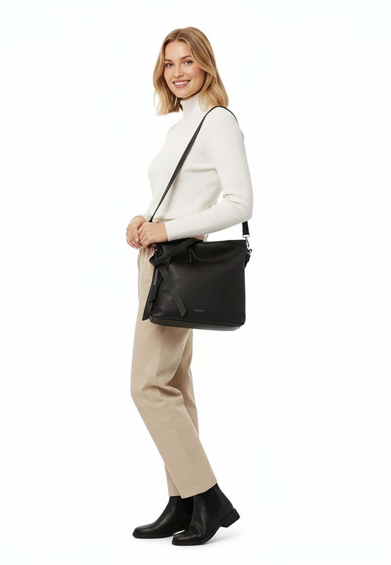 Donna con dolcevita bianco e pantaloni beige sorridente, che porta una borsa a tracolla in pelle nera e indossa stivaletti neri.