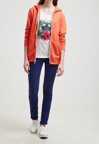 Orange zip-up hoodie med en lätt textur, lager på lager över en vit grafisk t-shirt med tropiska fåglar. Matchad med marinblå byxor och vita skor.