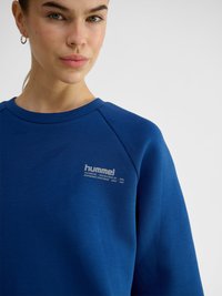 Giovane donna che indossa una felpa blu hummel con logo sul petto sinistro, che guarda leggermente verso la fotocamera su uno sfondo neutro.