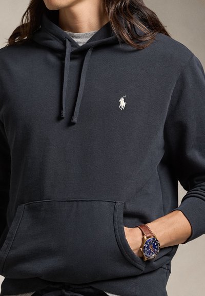 Camisola com capuz azul marinho escuro com bolso frontal, capuz com cordão e logótipo branco da Polo. Usada com um relógio de pulso com mostrador azul e pulseira de couro castanho.