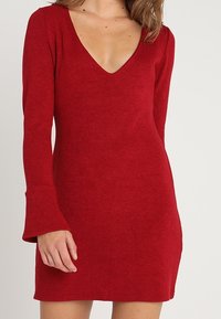 Röd stickad klänning med långa klockärmar och djup V-ringning. Tyget verkar mjukt och texturerat, vilket ger en figurnära siluett.