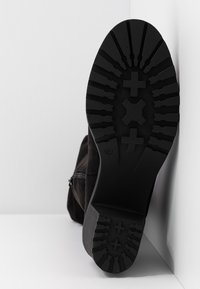 Semelle de botte noire avec des motifs de crantage profonds, incluant des croix et des nervures texturées, positionnée verticalement contre un mur d'angle blanc.