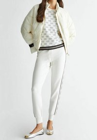 Giacca bianca imbottita, maglione a fantasia, pantaloni bianchi a vita alta con design laterale e ballerine con dettagli in oro metallico. Tessuti lisci ovunque.