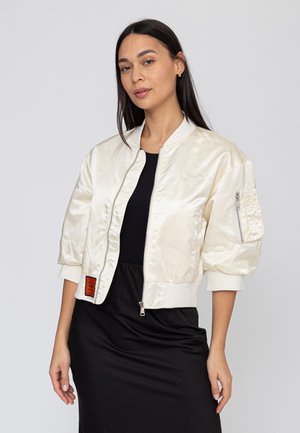 Femme aux longs cheveux noirs portant une veste bomber en satin crème sur une robe noire, debout devant un fond uni gris clair.