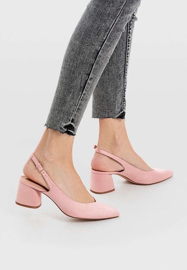 Personne portant un jean skinny gris et des chaussures slingback à talon bloc rose clair, debout sur un fond clair uni.