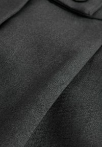 Schwarzer Stoff mit einer strukturierten, gewebten Oberfläche, die diagonale Falten aufweist und ein kleines Knopfdetail im oberen Bereich sichtbar ist.