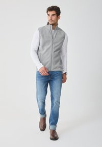Vest din fleece gri cu guler înalt, fermoar frontal și buzunare laterale, purtat peste o cămașă albă cu mâneci lungi și asortat cu blugi albaștri și bocanci maro.