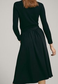 Femme portant un haut noir à manches longues et une jupe midi noire taille haute, vue de dos sur un fond uni.