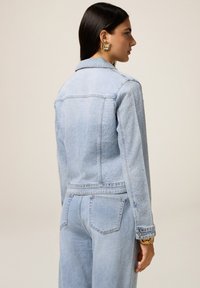 Veste en denim bleu clair avec des accents en strass, col classique, manches longues et deux poches arrière. Présente une coupe structurée et une texture délavée.