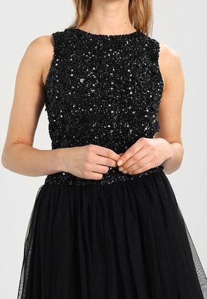 Black sleeveless dress with a sequined bodice and a flowing tulle skirt, worn by a person with hands clasped at the waist.
