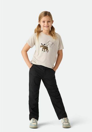 Ung pige med blondt hår i hestehaler iført beige t-shirt med elg- og bjergmotiv, sorte bukser og hvide sneakers, stående med hænderne i lommerne.