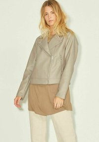 Giubbotto in pelle grigia per moto con design corto, tasche con zip e colletto asimmetrico, indossato sopra una maglia marrone e pantaloni chiari.