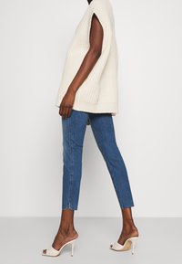 Jersey de punto grueso color crema sin mangas, jeans ajustados azules con un ligero desgate en los tobillos y tacones de punta abierta en color beige con un diseño acolchado.