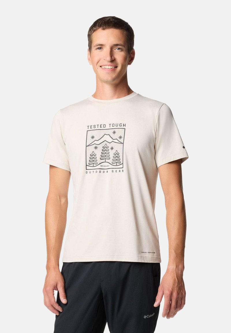 Lächelnder Mann, der ein hellbeiges T-Shirt mit Berg- und Baum-Motiv und schwarze Hose trägt, vor schlichtem hellem Hintergrund stehend.