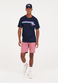 Camiseta de marina con rayas horizontales rojas y azules, combinada con pantalones cortos rosas. Las zapatillas son blancas con acentos negros. El modelo está de pie sobre un fondo plano.