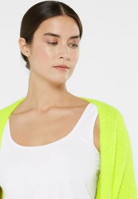 Neon green knitted cardigan with open front, contrasting with a white tank top. The texture is soft and fuzzy, enhancing the vibrant color.