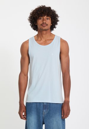 Homme aux cheveux bouclés portant un débardeur bleu clair et un jean bleu ample, debout devant un fond blanc.