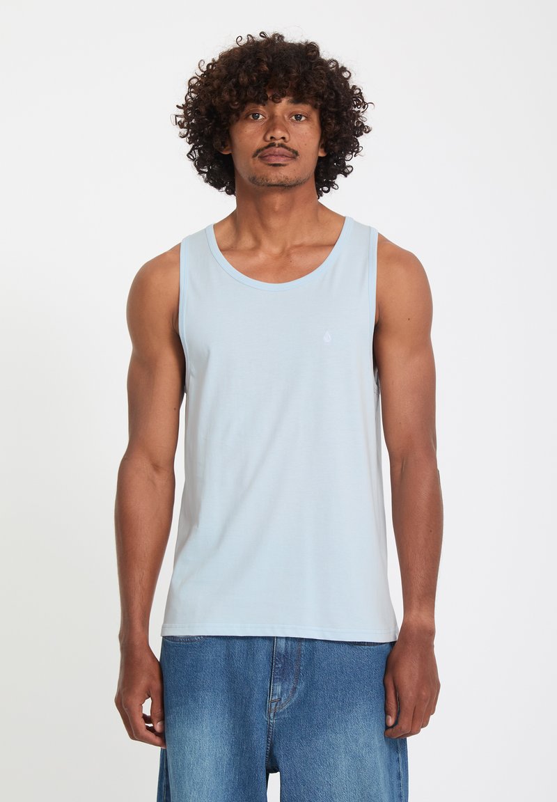 Homme aux cheveux bouclés portant un débardeur bleu clair et un jean bleu ample, debout devant un fond blanc.