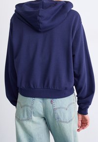 Hochtailliertes, navyblaues Kapuzenshirt aus weichem Stoff, ausgestattet mit einer Kapuze und gerippten Bündchen sowie Saum, kombiniert mit hellblauen Jeans mit Gesäßtaschen.