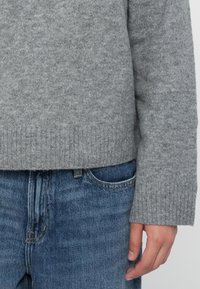 Persona che indossa un maglione a maglia grigia con polsini a costine e jeans di denim blu, con la mano appoggiata di lato.