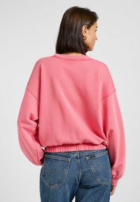 Roze sweatshirt met een relaxte pasvorm, geribbelde zoom en lange mouwen. Gemaakt van zacht materiaal. Gecombineerd met blauwe spijkerbroek.