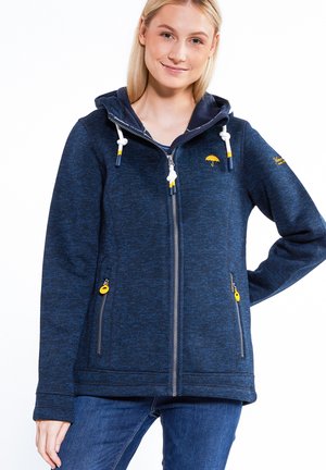 Frau trägt einen marineblauen Kapuzenpullover mit Reißverschluss und gelbem Regenschirm-Logo, kombiniert mit blauen Jeans, steht mit einer Hand in der Hüfte.