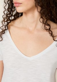 Bijela V-mašna majica izrađena od lagane, teksturirane tkanine. Ima kratke rukave i ležeran kroj, ističući suptilan, mekan završetak.