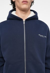 Sweat à capuche zippé bleu marine avec doublure en polaire, doté d'une fermeture éclair à l'avant, de deux poches avant et d'un logo brodé sur la poitrine gauche.