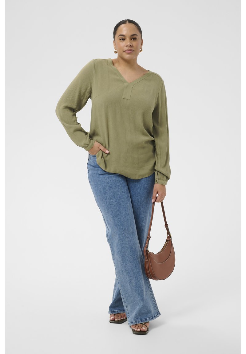 Femme portant un chemisier vert à manches longues et un jean bleu, tenant un sac à main marron, debout avec une main dans la poche et une jambe croisée.