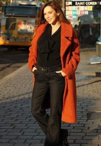 Lang oranje, gestructureerde jas over een zwarte blouse met een strikje, gecombineerd met donkere flared jeans. Staand op een stenen stoep in het zonlicht.