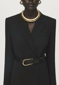 Zwarte op maat gemaakte blazer met een dubbelgebreide ontwerp, subtiele pocketdetails en een zwarte riem met een gouden gesp. Draagt een chunky gouden ketting.