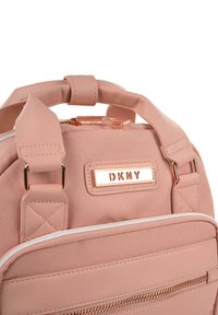 Mochila de tela rosa de DKNY con una textura suave, asas dobles y herrajes en oro rosa. Cuenta con un bolsillo con cremallera y una placa con el logotipo de la marca.