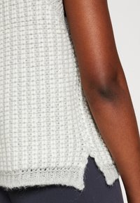 Braccio di pelle scura che indossa una maglietta senza maniche di maglia bianca con fessura laterale, abbinata a pantaloni blu navy, mostrato dalla vita al gomito.