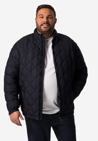 Mörk marinblå vadderad jacka med hög krage, har dragkedja fram och avslappnad passform, bärs över en vit T-shirt och kombineras med mörka jeans.