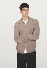 Ung man med kort mörkt hår, iklädd en beige zip-up tröja över en vit skjorta, med armarna i kors och tittar åt sidan.