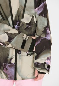 Eine gemusterte Jacke mit einem abstrakten Design in Grün-, Lila- und Beigetönen, mit schwarzen Reißverschlüssen und Klettverschlüssen an den Manschetten.