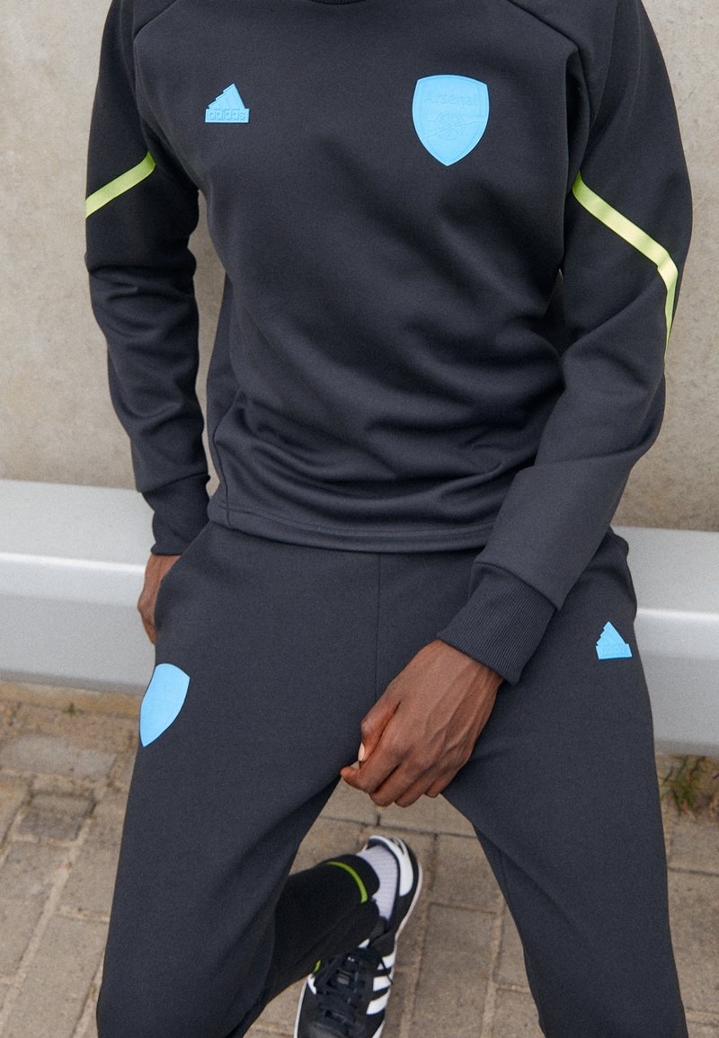 Tuta sportiva grigio scuro con dettagli azzurri e logo dell'Arsenal, caratterizzata da strisce verdi sulle maniche e polsini a costine sui pantaloni.