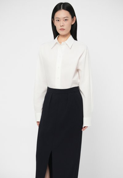 Γυναίκα με ίσια μαύρα μαλλιά που φοράει ένα άσπρο πουκάμισο με κουμπιά, χτενισμένο μέσα σε μια ψηλόμεση μαύρη φούστα με άνοιγμα μπροστά, που στέκεται μπροστά σε λευκό φόντο.