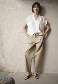 Vrouw met kort donker haar, draagt een losse witte v-hals blouse, beige wijde hoge taille broek en zwarte sandalen met hak, staand tegen een gestructureerde muur.