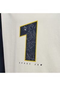 Vit T-shirt med en stor marinblå siffra "1" som är omgiven av gult, med tecknade figurer och texten "SPACE JAM" nedanför.