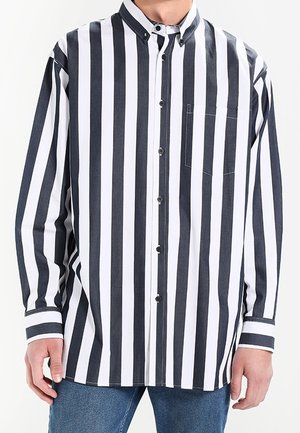 Mann, der ein schwarz-weiß gestreiftes Langarmhemd mit Knöpfen, einer Brusttasche und blaue Jeans trägt.
