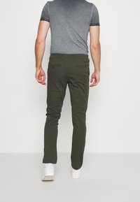 Man wearing dark green slim-fit pants, gray short-sleeve shirt, and white sneakers standing against a plain white background.