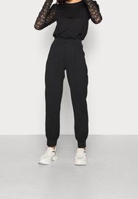 Czarne spodnie z wysokim stanem, z zakładkami, mankietami przy nogawkach i gładką fakturą, zestawione z czarną górą z koronkowymi, długimi rękawami.