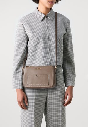 Borsa a tracolla in pelle marrone con tasca frontale, cuciture bianche e dettaglio logo dorato, abbinata a un outfit a motivo scozzese.